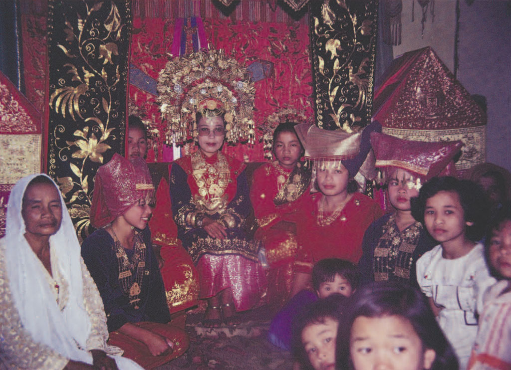 A group of people kneeling or sitting on rugs, a bride in the center in a massive, elaborate gold healdress, surrounding women in red fabric pronged headpieces.