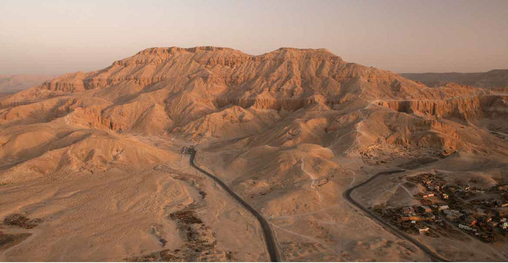Aerial view of the Valley of the Kings in Egypt, a winding road leading into cliffs, a town to the right at the foot.