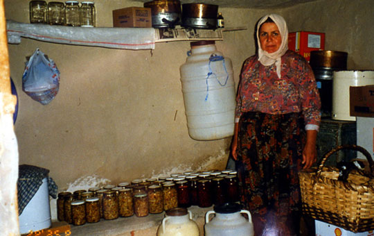 A woman in a windowless room with many jars of vegetables on a shelf.