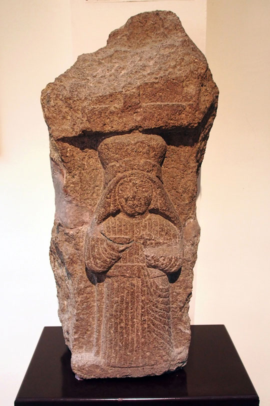 A stone carving of a person in lined robes and a large headpiece.