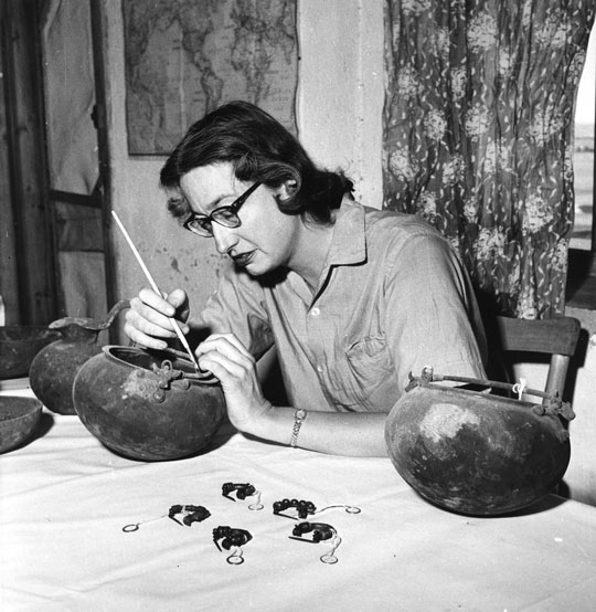 A woman at at able with several metal cauldrons, working delicately on one with a brush.
