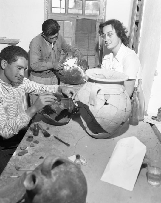 Two people sitting at work tables and working on fragmented pottery vessels with brushes while a woman looks on.