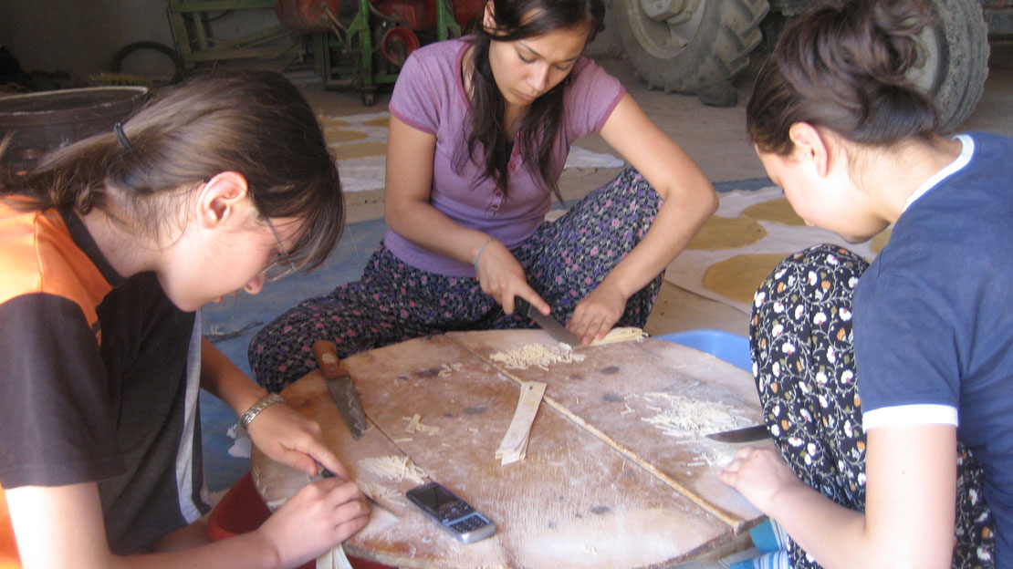 Three women sitting at a round table, working with knifes to cut small sticks.