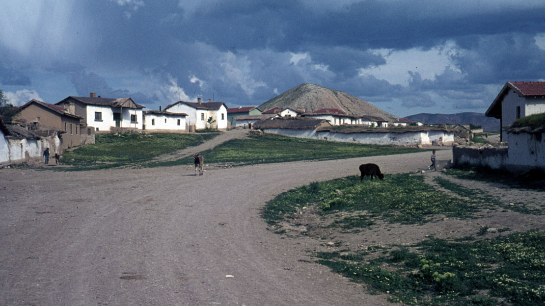 A road with houses lining the side, people walking along it. A massive mound in the distance.