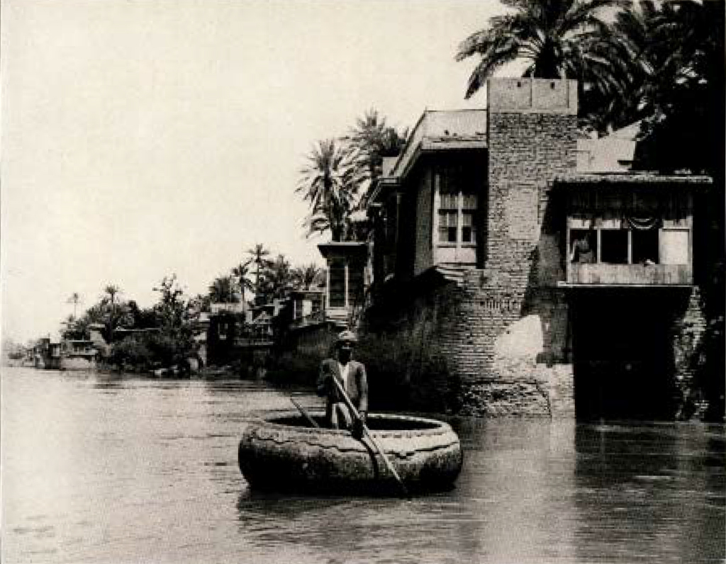 A man holding an oar standing in a circular boat on a river, along a row of houses and trees