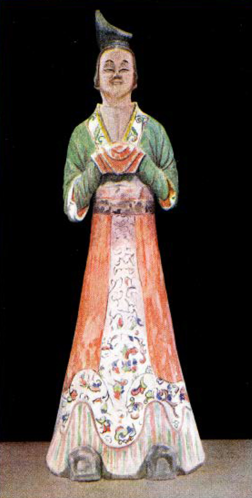 Painted pottery figure of a woman in a long red floral skirt with green long sleeve top, hands clasped together obscured by red pleated sleeves