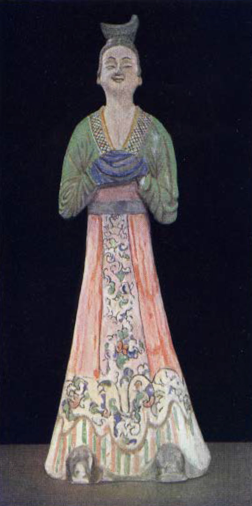 Painted pottery figure of a woman in a long red floral skirt with green long sleeve top, hands clasped together obscured by blue pleated sleeves