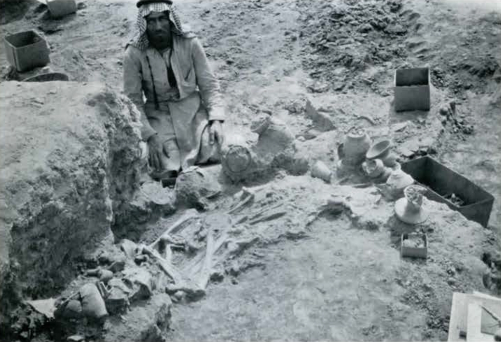 Remains of two people surrounded by pots and objects, a man kneeling next to the burial